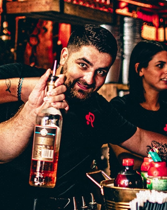 Barman Souriant Présentant une Bouteille d'Alcool au Bar Barman souriant tenant une bouteille d'alcool, ambiance festive de bar de nuit avec tatouages.
