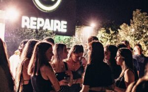 Groupe de jeunes fêtant devant l'enseigne néon REPAIRE lors d'une soirée animée.
